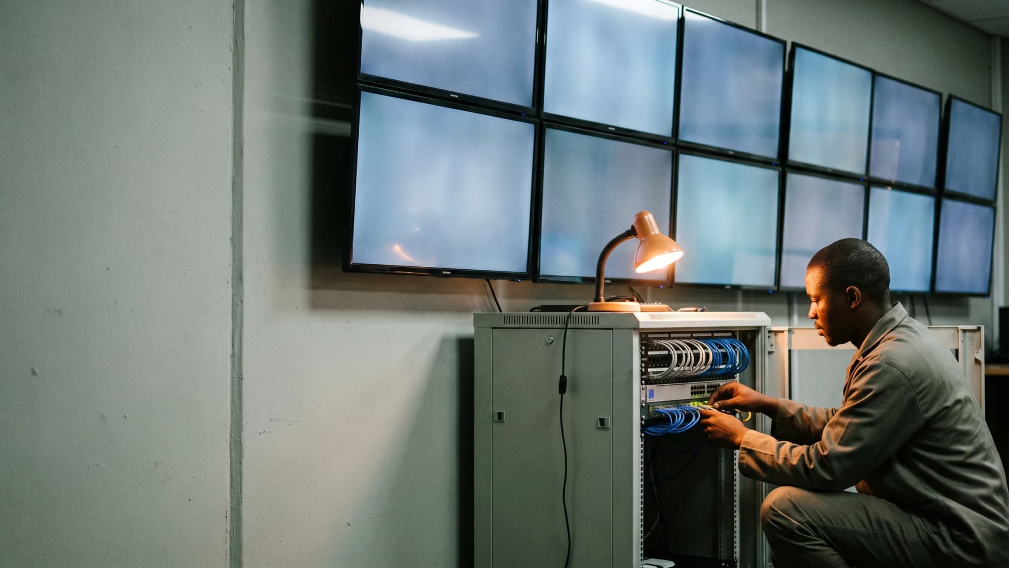 A technician wiring an equipment rack below a wall of modern flat-panel control room monitors