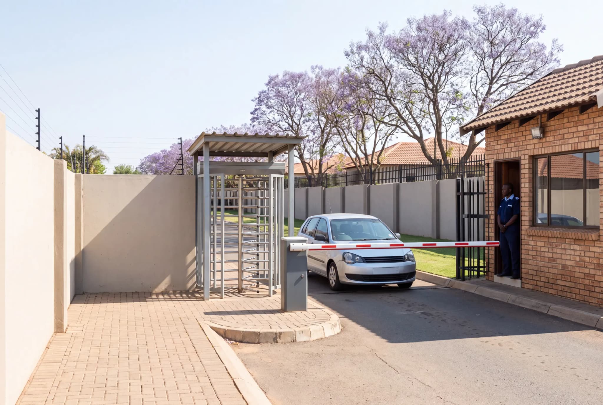 The entrance of a South African estate: a pedestrian turnstile, a car waiting at a lowered boom, and the guard in the doorway of the guard house